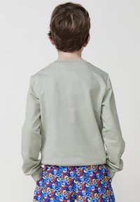 Camisa de manga larga verde claro con una textura suave, combinada con un pantalón corto azul que presenta un patrón de calavera pirata con tibias cruzadas en rojo y amarillo.