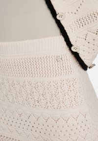 Nahaufnahme von cremefarbener Strickware mit Diamant- und Rautenmuster, versehen mit weißen Knöpfen und einem schwarzen Rand am Kleidungsstück.