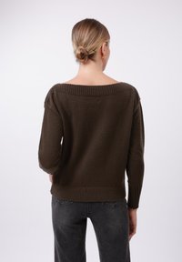 Brauner, gestrickter Pullover mit weitem, geripptem Kragen und geripptem Saum, im lässigen Schnitt mit langen Ärmeln, getragen über dunkelgrauen Jeans.