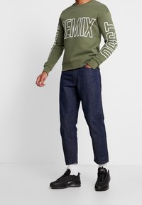Felpa verde oliva con testo bianco "REMIX" sul davanti e sulle maniche, abbinata a jeans blu scuro e sneakers nere.