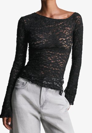Femme portant un haut en dentelle noire cintré à manches longues, associé à un jean taille haute gris clair, main dans la poche.