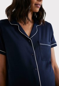 Granatowa koszula piżamowa z miękkiego materiału, z białymi lamówkami wzdłuż kołnierza, listwy guzikowej i kieszeni. Krótkie rękawy i detal pojedynczej kieszeni na piersi.