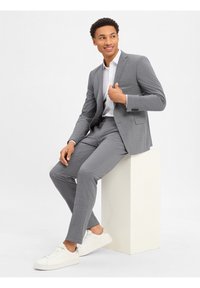 Lichtgrijs pak met een slim fit, gestructureerde stof, wit overhemd en witte sneakers. Onderwerp zit op een voetstuk met een neutrale achtergrond.