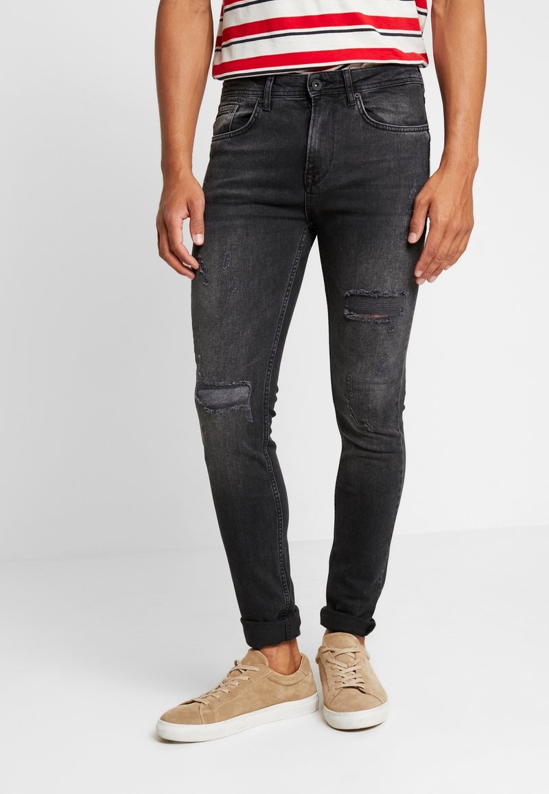 Jeans ajustados de color gris oscuro con detalles desgastados en las rodillas, cinco bolsillos y costuras sutiles. Combinados con zapatillas de gamuza beige.