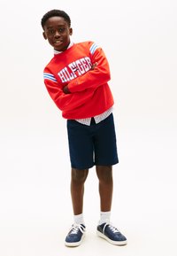 Chlapec v oranžovej mikine s nápisom „HILFIGER“, pruhovanom tričku, námornícky modrých šortkách, bielych ponožkách a modrých teniskách pózuje so založenými rukami.