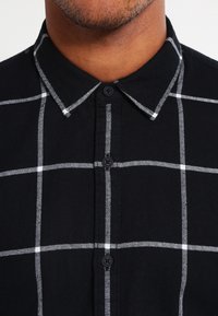 Primo piano di una persona che indossa una camicia nera abbottonata con un motivo a quadratini bianchi e colletto a punta.