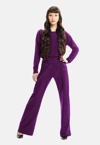 Lila tvådelad outfit bestående av en långärmad topp och vida byxor. Slät tyg med avslappnad passform och enkel design. Svarta högklackade skor.