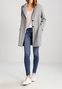 Abrigo gris texturizado con solapas anchas, cierre de doble botonadura y bolsillos laterales, usado sobre una blusa clara y jeans ajustados azules, combinado con zapatillas blancas.