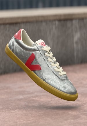 Sneaker argentato con logo rosso a V, lacci crema e suola in gomma, sospeso sopra una superficie di cemento con sfondo grigio sfocato.