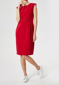 Rotes ärmelloses Kleid mit tailliertem Schnitt und Schleifendetail, knielang, aus glattem Stoff; kombiniert mit weißen Sneakern.