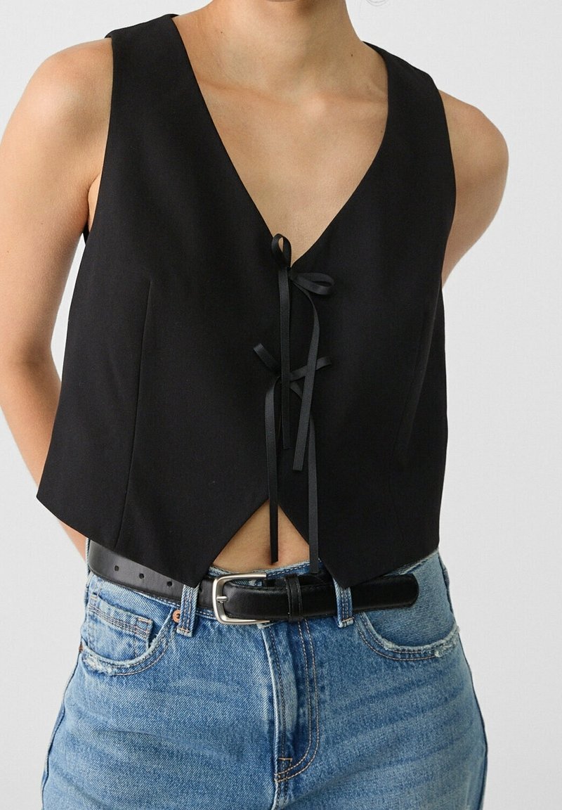 Schwarze, kurz geschnittene Weste mit tiefem V-Ausschnitt, ausgestattet mit einer vorderen Knotenbindung und einem scharfen Saum. Kombiniert mit hoch taillierten blauen Jeans und einem schwarzen Gürtel.