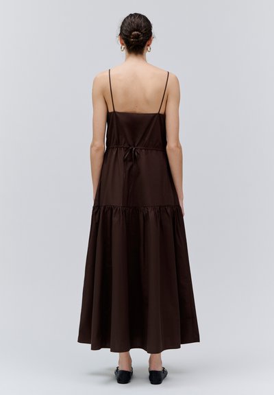 Femme aux cheveux attachés en chignon, portant une robe maxi marron foncé sans manches avec fines bretelles et des chaussures plates noires, debout devant un fond uni.