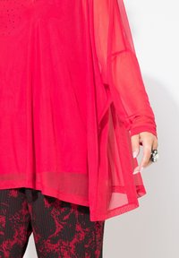 Rote, durchsichtige Tunika mit fließendem Design und langen Ärmeln, kombiniert mit strukturierten schwarzen und roten gemusterten Hosen. Auffälliger Ring an der Hand.