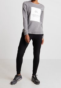 Sweatshirt gris avec logo blanc, porté avec un pantalon noir et des chaussures de sport noires. Coupe ajustée, texture moyenne.