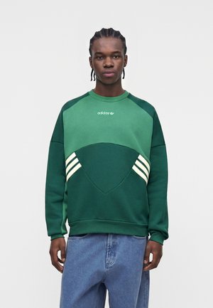 Giovane uomo con capelli intrecciati che indossa una felpa Adidas verde bicolore con strisce bianche e jeans blu, in piedi davanti a uno sfondo semplice.