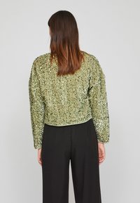 Błyszcząca zielona krótka kurtka z cekinami, z długimi rękawami i gładką podszewką, noszona na czarnych spodniach, które mają teksturowaną powierzchnię.