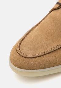 Zapato de ante marrón con punta redondeada, detalle de costura visible a lo largo del borde y suela de goma de color claro.