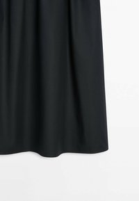 Jupe en tissu noire suspendue sur un fond blanc, montrant la partie inférieure avec des plis doux et une texture lisse.