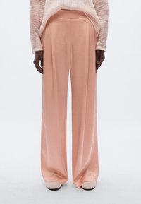 Pantalon large rose à texture lisse, avec plis sur le devant et taille haute, associé à des chaussures de couleur claire.