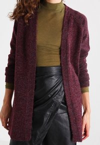 Bordeauxstickad cardigan med långa ärmar och öppen framsida, lagd över en olivgrön polotröja. Svart fusk läder omslagskjol fulländar looken.