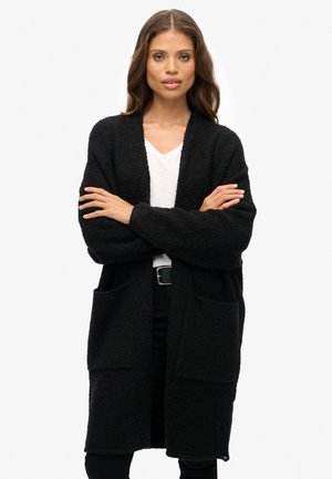 Frau mit langen Haaren, die eine schwarze, strukturierte Strickjacke über einem weißen Oberteil und schwarzen Hosen trägt, mit verschränkten Armen vor einem weißen Hintergrund steht.