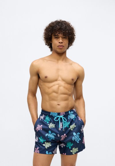 Σορτς κολύμβησης σε ναυτικό μπλε με πολύχρωμα μοτίβα χελώνας. Διαθέτει ελαστική μέση με τιράντα σε τυρκουάζ και πλαϊνές τσέπες.
