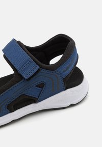 Blå och svarta sportsandaler med en övre del i tyg, justerbart rem och vit gummisula med texturerad detalj.