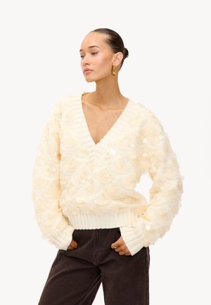 Suéter color crema con escote en V, texturizado con escamas o pétalos superpuestos, puños y dobladillo acanalados. Combinado con pantalones marrón oscuro.