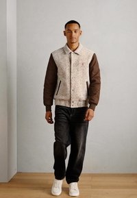 Giacca trapuntata con motivo a spina di pesce in beige e marrone, con maniche marroni scure, tasche laterali e chiusura a pulsante. Abbinata a jeans scuri e sneakers bianche.