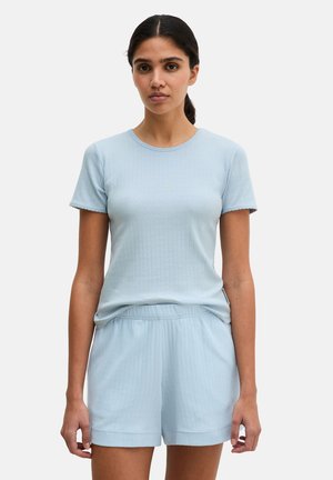 Vrouw staand, gekleed in een lichtblauw geribd shirt met korte mouwen en bijpassende shorts met elastische taille, tegen een effen achtergrond.