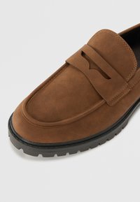 Mocassins en suede marron avec un accent de bande surélevée, bout arrondi et semelle en caoutchouc texturée. Présente des coutures visibles et une finition lisse.