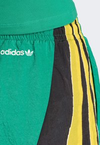 Gröna och svarta shorts med gula och svarta vertikala ränder, utrustade med elastisk midja och vit Adidas-logotyp. Texturerat tyg.