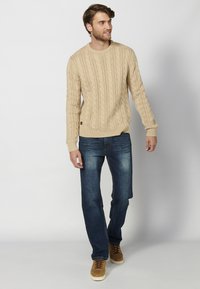 Maglione beige a treccia, collo rotondo, polsini e orlo a coste, abbinato a jeans blu in denim, sneakers marroni e uno sfondo neutro.