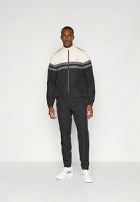 Melna un bēša sporta tērpa komplekts ar horizontālām pelēkām svītrām uz krūtīm. Viegla auduma jaka ar rāvējslēdzēju un sašaurinātas bikses ar kabatām.