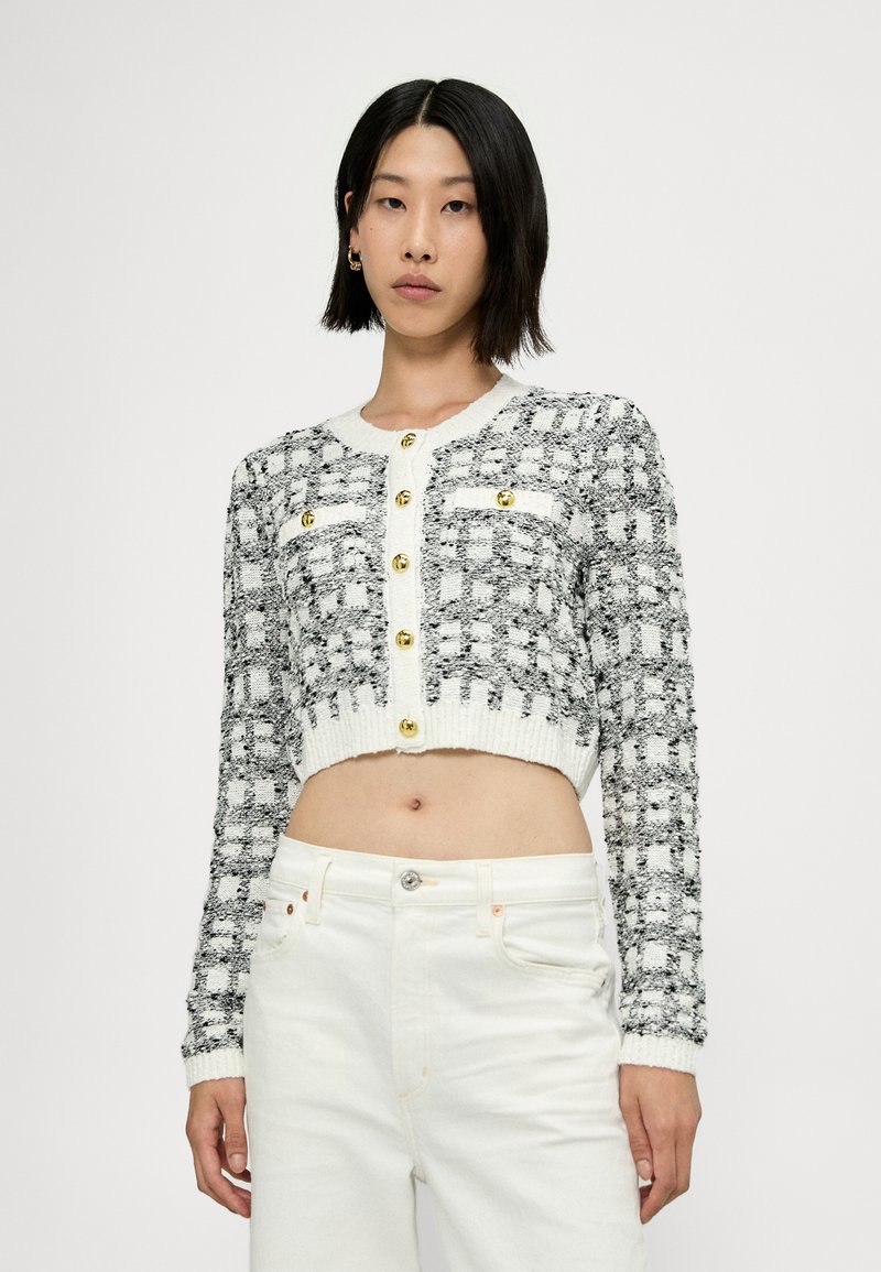 Cardigan court gris et blanc avec une texture tissée, comportant des boutons dorés et deux poches avant. Associé à un pantalon blanc taille haute.