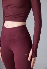 Kirsipunane pikas varrukateta crop top, mis on valmistatud ribakanga materjalist, koos kõrge vöökohaga retuusidega; mõlemal on sujuv tekstuur ja diskreetne logodetail.
