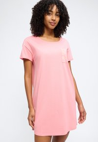 Γυναίκα με μπούκλες, φορώντας ένα φαρδύ ροζ φόρεμα με κοντά μανίκια και μια μικρή τσέπη στο στήθος με λουλουδάτο μοτίβο, σε απλό φόντο.