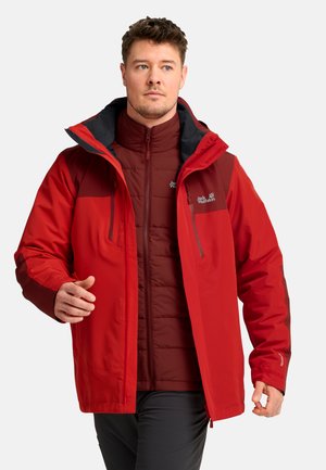 Uomo che indossa giacche outdoor sovrapposte rosse e rosso scuro con loghi, che posa davanti a uno sfondo chiaro uniforme.