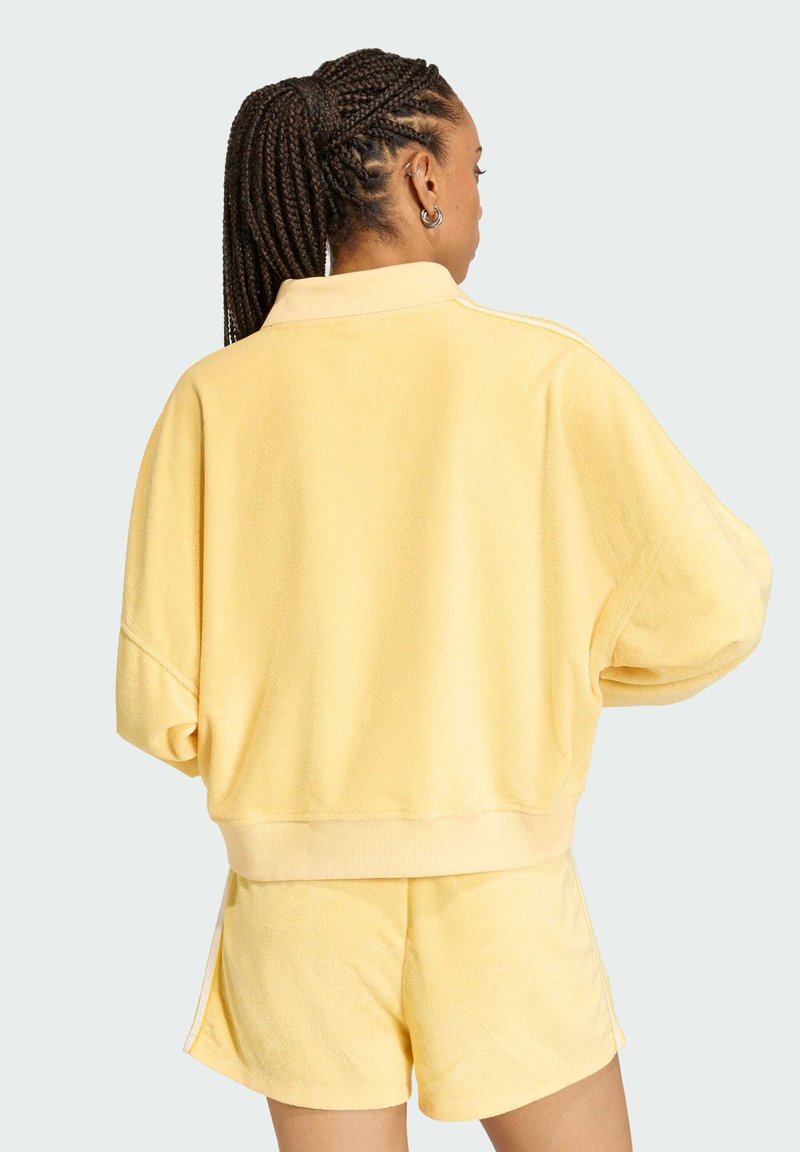 Tuta di felpa giallo composto da una giacca corta e ampia con colletto e pantaloni corti coordinati, entrambi caratterizzati da una texture morbida e strisce laterali.