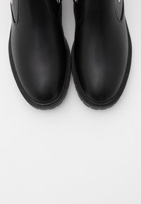 Černé kotníkové boty vyrobené z hladkého syntetického materiálu, s kulatou špičkou, texturovanou gumovou podešví a dekorativními kovovými akcenty.