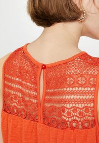 Pomarańczowa koszulka bez rękawów z ażurowym tyłem o teksturowanym wzorze w okręgi oraz małym zapięciem na guziczek przy szyi. Miękka tkanina.