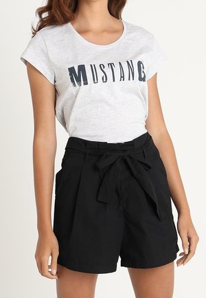 Woman wearing a light grey "MUSTANG" text t-shirt tucked into high-waisted black shorts with a tied fabric belt.