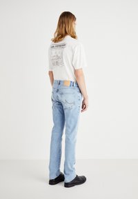 Vit t-shirt med grafik från ett kafé på baksidan, i kombination med ljusblå denimjeans som har en beige patch vid midjan och bakfickorna.