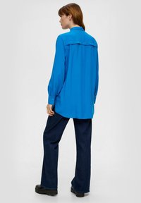 Blauwe lange mouwen shirt met knoopmanchetten, rugpasdetail en een relaxte fit, gecombineerd met donkere spijkerflarebroeken en zwarte schoenen.