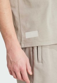 Jasnobeżowa koszulka z teksturowanym wykończeniem, bocznymi szwami i dyskretną naszywką z logo blisko dołu, zestawiona z pasującymi beżowymi spodniami.