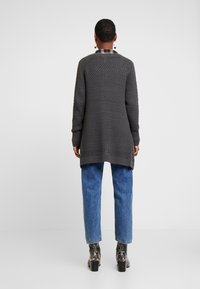 Cardigan grigio lavorato a maglia con un motivo testurizzato, maniche lunghe, spacchi laterali e polsini a costine, abbinato a jeans blu e stivali neri con tacco.