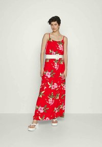 Mulher a usar um vestido maxi sem mangas vermelho com flores, um amplo cinto branco e sandálias de plataforma brancas, em pé contra um fundo claro e liso.