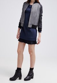 Woman wearing navy dress with black pleated hem, gray and black bomber jacket, black ankle boots, and black socks against white background.