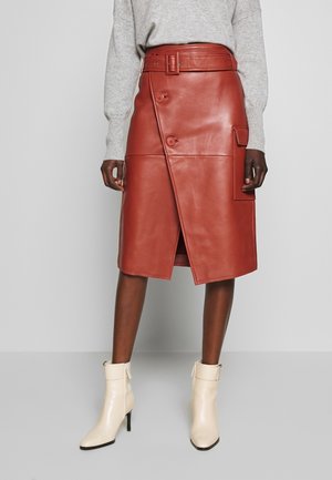 Model iført et knælangt rustbrunt læder omslagsnederdel med knapper og bælte, kombineret med en grå sweater og cremefarvede ankelstøvler.