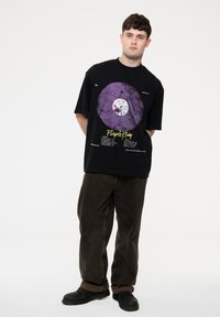 Black short-sleeve t-shirt featuring a purple vinyl record graphic and text; paired with loose brown corduroy pants and black shoes.